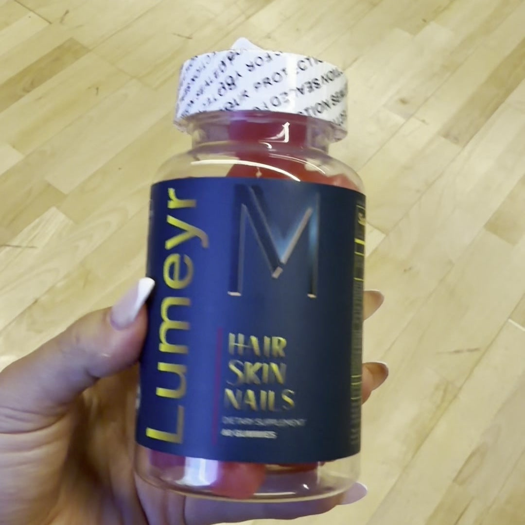 Hand holding a bottle of Lumeyr Hair Skin Nails gummies with the gold Lumeyr wordmark and M monogram on a navy label