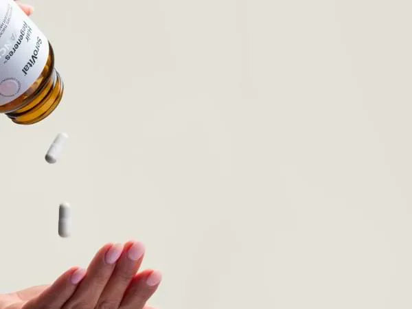 Hand holding a bottle of supplements with pills falling onto a light surface